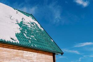 Snow lies on the green tiled roof of a wooden cottage against the blue sky photo