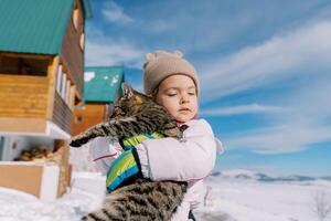 pequeño niña abrazos un atigrado gato mientras en pie en el nieve cerca un de madera cabaña foto