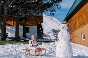 pequeño niña se sienta en un trineo cerca un monigote de nieve siguiente a un de madera casa foto