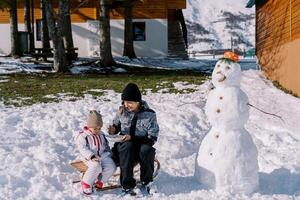 mamá alimenta un pequeño niña desde un cuchara, sentado en un trineo cerca un monigote de nieve cerca un cabaña foto