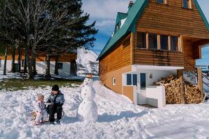 Mom feeds little girl from a spoon sitting on a sled in the courtyard of a wooden chalet photo