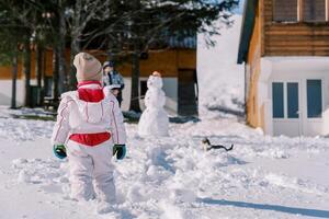 Small child looks at his mother standing near a snowman next to a walking cat in the yard. Back view photo