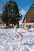 Small child pulls a sleigh on a rope past a wooden chalet, looking back at the snowman photo