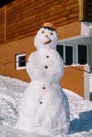 Snowman with a saucepan with red polka dots on his head stands near a wooden house photo