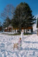 pequeño niña soportes cerca un trineo siguiente a un monigote de nieve en el yarda de un de madera casa foto