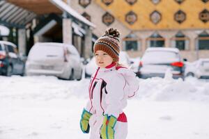 pequeño niña soportes oblicuo en el nieve cerca carros estacionado cerca el hotel foto