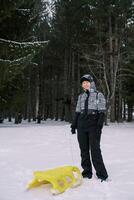 Smiling young woman stands with a yellow sled in the snow near a coniferous forest photo