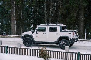 White Hummer H2 pickup truck drives under snowfall along a snow-covered road along a coniferous forest. Side view photo