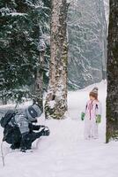 pequeño niña soportes en el bosque y relojes su madre haciendo un monigote de nieve foto