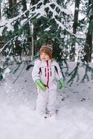 pequeño niña en un esquí traje soportes debajo un cubierto de nieve Navidad árbol en el bosque foto