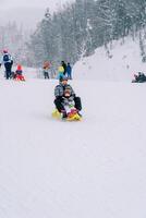 sonriente madre con un pequeño niño va abajo el Pendiente de un Nevado montaña en un trineo foto