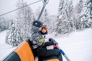 Smiling mother with a little girl in her arms rides a chairlift up a snowy hill photo