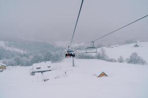 Skiers ride a chairlift up the mountain to snow-covered cottages among a coniferous forest photo