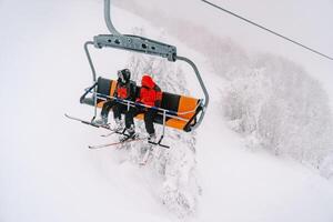 Skiers ride a chairlift up a foggy mountain above a snowy forest photo