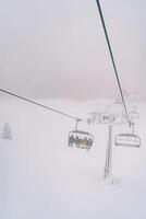 turistas en vistoso esquí trajes paseo cuesta arriba mediante el niebla en un telesilla encima un Nevado bosque foto