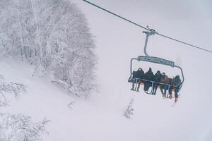 Tourists in ski equipment on skis climb up the mountain on a chairlift above the snowy trees photo