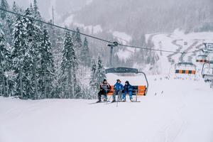Skiers in ski suits ride on a ski lift along a snowy wooded mountain slope photo
