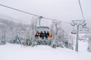 Skiers in bright suits ride on a chairlift over a snowy forest up in the mountains photo