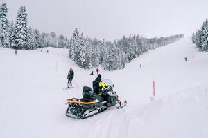 Man on a snowmobile rides past skiers descending a snowy slope. Back view photo