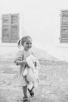 Little smiling girl walks hugging a toy rabbit near the house. Black and white photo