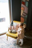 Little girl stands near a chair with a sleeping tabby cat and looks away photo