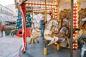 pequeño niña paseos un juguete caballo en un carrusel a un justa cerca un decorado Navidad árbol cerca un antiguo casa foto