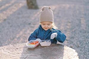 Little smiling girl scoops porridge with a spoon from a lunchbox while standing behind a tree stump photo