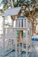 pequeño niña soportes en el plataforma de un juguete de madera casa en el patio de juegos, propensión en el pretil y mirando dentro el distancia foto