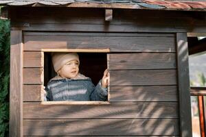 pequeño niña mira fuera de el ventana de un juguete de madera casa en el patio de recreo foto