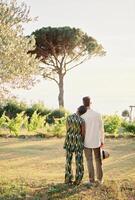 Woman rested her head on man shoulder while standing with him in a green garden. Back view photo