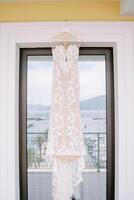 Lace wedding dress with straps hanging on a hanger on the balcony with the reflection of the sea and mountains in the glass photo