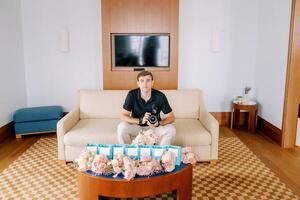 Photographer with a camera sits on a sofa in front of a table with small bouquets of flowers and blue gift bags photo