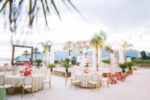 Festive tables with colorful bouquets of flowers stand around a round stage on the seashore against the background of mountains photo