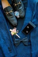 Black men shoes stand on a blue suit next to a watch and boutonniere photo