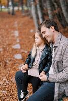 sonriente papá inclinado su frente en contra el cabeza de un pequeño niña sentado en su regazo en un banco en el otoño bosque foto