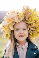 Smiling little girl in a wreath of autumn leaves stands in a meadow. Portrait photo