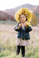 Little girl in a wreath of autumn leaves looks at a yellow leaf in her hand while standing on the lawn photo