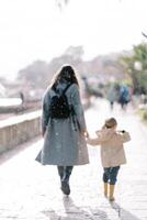 Mom and little girl are walking holding hands under the snowfall on the embankment. Back view photo