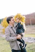 Dad with a little girl in a wreath of yellow leaves in his arms stands in a clearing photo