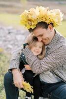 Smiling dad in a wreath of yellow leaves hugs a little girl sitting on his lap photo