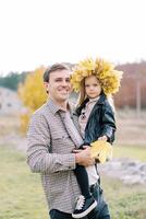 sonriente papá participación en su brazos un pequeño niña en un guirnalda de otoño hojas con un amarillo hoja en su mano foto