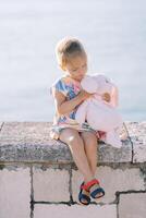 Little girl examines a plush rabbit in her hands while sitting on a fence by the sea photo