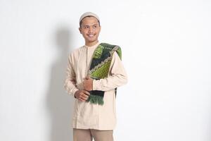Portrait of religious Asian muslim man in koko shirt with prayer mat standing with cool pose and holding his sajadah. Isolated image on white background photo