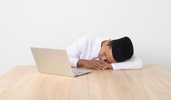 Portrait of excited Asian muslim man in koko shirt with skullcap working during fasting on ramadan month, feeling sleepy, sleeping at his table with laptop. Isolated image on white background photo