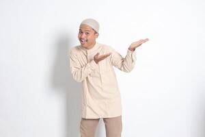 Portrait of shocked Asian muslim man in koko shirt with skullcap showing product and pointing with his hand and finger to the side. Advertising concept. Isolated image on white background photo