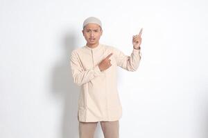 Portrait of shocked Asian muslim man in koko shirt with skullcap showing product and pointing with his hand and finger to the side. Advertising concept. Isolated image on white background photo