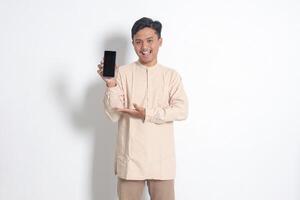 Portrait of young excited Asian muslim man in koko shirt showing blank screen mobile phone mockup while pointing and presenting product. Social media concept. Isolated image on white background photo