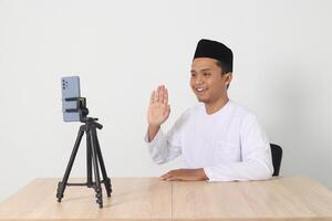 Portrait of excited Asian muslim influencer in koko shirt with skullcap saying hi, waving hand to phone camera during live streaming session. Isolated image on white background photo