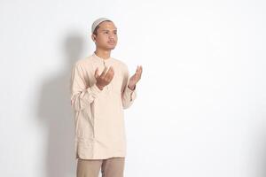 retrato de religioso asiático musulmán hombre en koko camisa con casquete Orando formalmente con su manos aumentó. devoto fe concepto. aislado imagen en blanco antecedentes foto