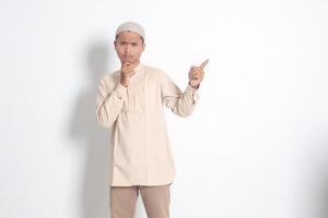 Portrait of shocked Asian muslim man in koko shirt with skullcap showing product and pointing with his hand and finger to the side. Advertising concept. Isolated image on white background photo